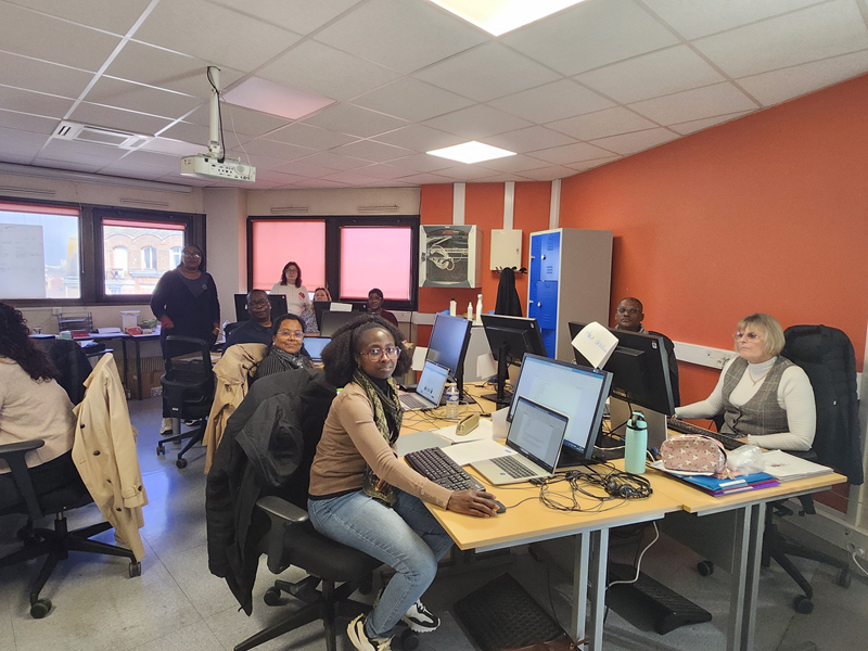 Plusieurs personnes sont assises autour de tables dans une salle de formation ou de bureau. Chacune travaille sur un ordinateur portable ou fixe. Certaines regardent vers l’appareil photo. La salle est éclairée, avec des murs orange et des fenêtres au fond. 