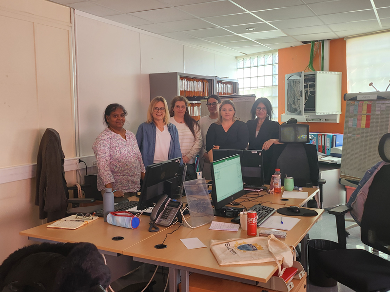 Image 1 :
Un groupe de six femmes pose dans un bureau. Elles sont debout derrière un bureau avec plusieurs ordinateurs, téléphones et objets du quotidien (bouteilles, carnet). La pièce est lumineuse, avec des étagères de dossiers à l’arrière. 