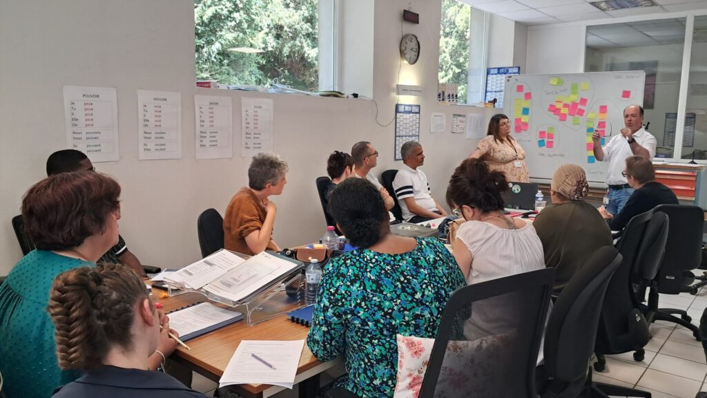 Réunion de travail avec plusieurs participants autour d’une table, pendant une présentation animée devant un tableau rempli de notes colorées.